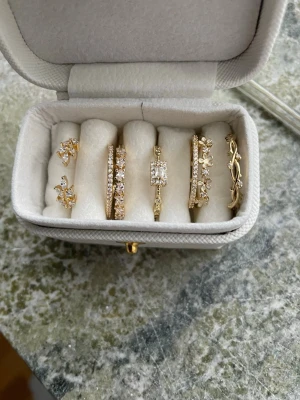 5 ringar justerbara & guld pläterade i guld 18k med stenar - Säljer ett set med fem snygga ringar i copper pläterade i guld 18k, dekorerade med gnistrande stenar och detaljerade mönster. Några ringar har blommiga inslag och andra är prydda med små kristaller. Måttet på alla 1,55-1,95 cm diameter. 4 har öppning och kan justeras i storleken, en har inte öppning och har i mått 1,6 cm diameter.Perfekt för att mixa och matcha till olika outfits.