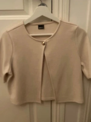 Beige kort kofta med knapp - Säljer en beige kort kofta från Zara med rund halsringning och en dekorativ guldknapp framtill. Koftan har korta ärmar och är tillverkad i ett mjukt, stickat material som ger en clean och stilren look.