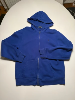 Blå hoodie från Polo Ralph Lauren - Klassisk blå hoodie från Polo Ralph Lauren med dragkedja framtill och huva. Hoodien har långa ärmar, två fickor fram och en liten broderad logga på bröstet. Perfekt för en chill och avslappnad stil.