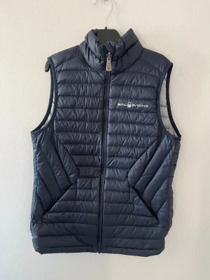 Mörkblå dunväst från Sail Racing - Snygg mörkblå dunväst från Sail Racing med hög krage och dragkedja framtill. Västen har två stora fickor på sidorna och diskret logga på bröstet. Perfekt för lager-på-lager och håller dig varm under kyliga dagar.