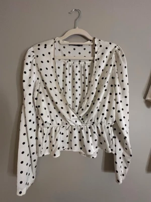 Vit prickig blus med omlott - Snygg vit blus med svarta prickar och omlott-skärning framtill. Blusen har lång ärm, markerad midja med volangkant och djup v-ringning. Perfekt för dig som gillar trendiga och lekfulla mönster. Materialet känns lätt och luftigt.