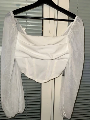 Vit off-shoulder topp med puffärmar - Trendig vit off-shoulder topp med puffiga, transparenta ärmar och draperad front. Korta längden och markerad midja ger en snygg siluett. Perfekt för att skapa en stilren och modern look.