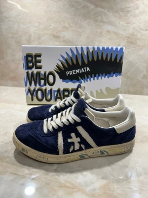 Premiata skor S 40 - Snygga sneakers från Premiata i mörkblå mocka med vita detaljer och snörning. Skorna har en låg siluett och en vit sula med tryck. Perfekta för dig som gillar en stilren men sportig look. Skick 9/10