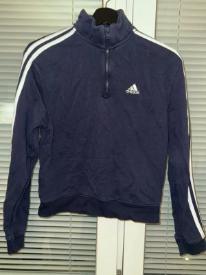 Mörkblå Adidas hoodie med dragkedja - Mörkblå hoodie från Adidas med halv dragkedja och klassiska vita ränder längs ärmarna. Tröjan har hög krage, muddar vid ärmslut och nederkant samt Adidas-logga på bröstet. Perfekt för en sportig och avslappnad stil.