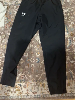 Svarta träningsbyxor Under Armour - Säljer ett par svarta träningsbyxor från Under Armour med resår i midjan och logga på vänster lår. Byxorna är raka i modellen och tillverkade i ett lätt och funktionellt material som passar perfekt till träning.