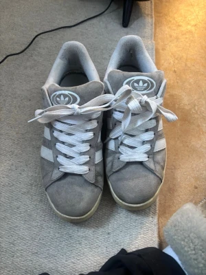 Adidas Campus grå sneakers - Gråa Adidas Campus sneakers i mocka med vita klassiska tre ränder på sidorna och vit sula. Skorna har vita skosnören och Adidas logga på plösen. Perfekt för dig som gillar en clean och tidlös stil.