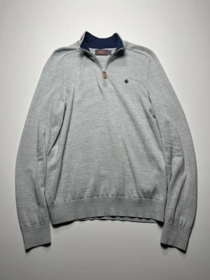 morris quarter zip - Ljusgrå stickad tröja från Morris med halv dragkedja och mörkblå broderad lilja på bröstet. Tröjan har ribbade muddar och krage med mörkblå insida. Perfekt för lager-på-lager och stilren look.