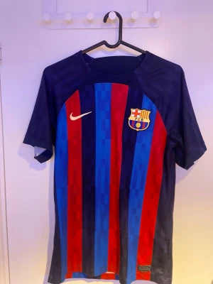 FC Barcelona matchtröja Nike 22/23 - Säljer en officiell FC Barcelona matchtröja från Nike med klassiska blå och röda vertikala ränder. Tröjan har klubbmärket på bröstet och Nike-logga i vitt. Tillverkad i lätt och ventilerande material, perfekt för fotbollsträning eller att bära på läktaren. Denna tröja är köpt utan reklam eller namn/nummer på baksidan