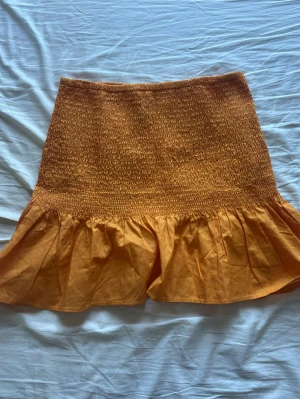 Orange volang tubtopp/kjol - Superfin orange kortkjol/bandeutopp med smockad midja och bred volang nertill. Kjolen är gjord i bomull 