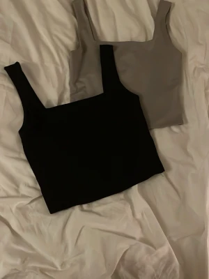 Två croppade linnetoppar - Två stilrena croppade linnetoppar i svart och grå. Båda har breda axelband och är ärmlösa, de slutar ungefär vid navlan!