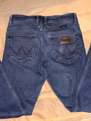 Blå Wrangler jeans med läderpatch - Klassiska blå jeans från Wrangler med raka ben och tydliga W-detaljer på bakfickorna. Jeansen har en brun läderpatch med Wrangler-logga och är tillverkade i slitstark denim. Perfekta för en avslappnad och tidlös stil.