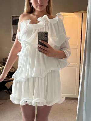 STUDENT klänning volanger - Säljer en vit kortklänning med stora volanger och omlott-design. Klänningen har en offshoulder-stil och är tillverkad i ett lätt, plisserat material som ger ett luftigt och trendigt intryck. Perfekt för dig som vill sticka ut med en unik look, och mer än student klänning, väldigt bekväm och man behöver ingen bh om man har mindre byst, storlek L men än s-m mer.