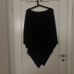Svart stickad poncho från KappAhl - Snygg svart stickad poncho från KappAhl med asymmetrisk form och lös passform. Perfekt att slänga över en tröja för en cool och avslappnad look. Enkel att matcha med olika outfits och passar året runt.