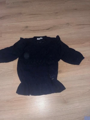 Mörkblå blus med volanger - Mörkblå stickad tröja från Holly & Whyte med fina hålmönster, volangdetaljer på axlarna och en bred volang nertill. Tröjan har rund halsringning och trekvartsärmar. Perfekt för dig som gillar en söt och trendig look.  