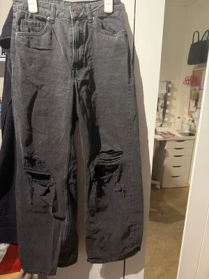 Svarta ripped jeans med raka ben - Säljer ett par svarta jeans med raka ben och stora slitningar över knäna. Jeansen har klassisk femficksmodell, hög midja och en avslappnad passform. Materialet är denim i bomull. Perfekta för en cool och avslappnad stil.