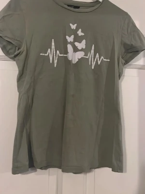 Grön t-shirt med fjärilar och hjärtslag - Snygg grön t-shirt med vitt tryck av fjärilar och hjärtslag på bröstet. Klassisk rund halsringning och korta ärmar. Perfekt för dig som gillar enkla men coola motiv. Tillverkad i mjuk bomull för skön känsla hela dagen. Som ni ser på bilden finns det lite fläckar som kanske går bort i tvätt, är inte helt säker🥰