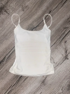 Vit linnetopp med smala axelband - En enkel och stilren vit linnetopp med tunna axelband och rak passform. Perfekt att matcha med jeans eller kjol för en clean look. Materialet känns mjukt och lätt mot huden.