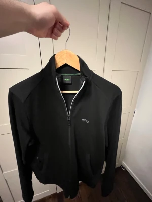 Svart ziptröja från BOSS - Snygg svart ziptröja från BOSS. Tröjan har hög krage, dragkedja framtill och är tillverkad i ett mjukt material