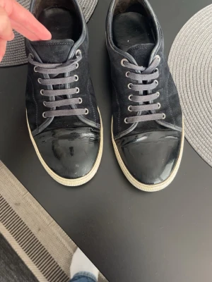 Lanvins - Säljer ett par svarta sneakers med coola lackade tår och grå skosnören. Skorna har vit sula och metallfärgade öljetter. Perfekta för dig som vill ha en stilren men ändå unik look.