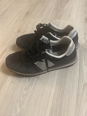 Svarta New Balance sneakers - Snygga svarta sneakers från New Balance med klassisk silvrig N-logga på sidan. Skorna har låga skaft, svarta snören och detaljer i vitt och grått. Ovandelen är i mesh och mocka för en sportig look. Perfekta för dig som gillar streetwear och bekväma skor.