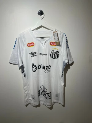 Vit Santos FC matchtröja från Umbro - Snygg vit matchtröja från Santos FC med flera sponsorloggor och klubbmärke på bröstet. Tröjan har rund hals, korta ärmar och är tillverkad i ett lätt och ventilerande material som passar perfekt för fotbollsträning eller match.