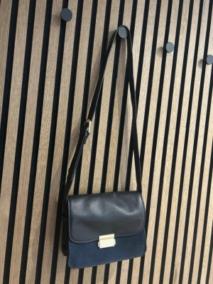 Svart axelväska i läder och mocka - Snygg svart axelväska med justerbar rem, kombinerar slätt läder på locket och mjuk mocka på väskans framsida. Stängs med ett stilrent guldfärgat spänne. Perfekt storlek för det viktigaste och ger en clean look till din outfit.