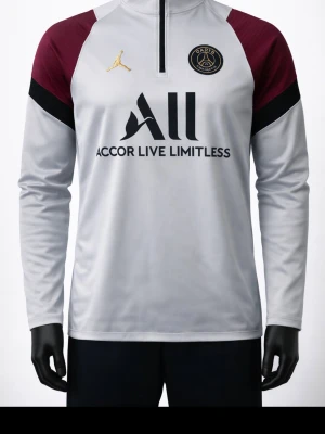 PSG långärmad träningströja - Snygg långärmad träningströja från PSG i samarbete med Jordan. Tröjan är vit med vinröda axlar och svarta detaljer, har halv dragkedja och klubbmärke på bröstet. Tillverkad i funktionsmaterial som passar perfekt till träning.