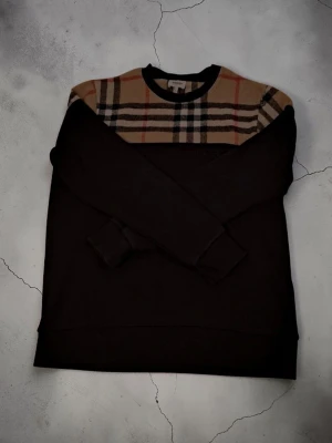 Svart sweatshirt med Burberry-rutor - Säljer en svart sweatshirt från Burberry med klassiskt rutmönster i beige, rött och svart över axlar och bröst. Tröjan har rund halsringning och långa ärmar. Materialet känns mjukt och bekvämt, perfekt för en stilren och trendig look.