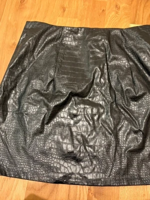 Svart kjol i skinnimitation från SHEIN - Snygg svart kjol från SHEIN i skinnimitation med croco-mönster. Kjolen har en kort passform och är perfekt för dig som vill sticka ut med en trendig look. Materialet är syntet och ytan har en cool struktur som påminner om krokodilskinn.