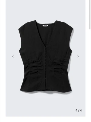 Svart v-ringad blus från Weekday - Stilren svart blus från Weekday med v-ringning och knappar framtill. Modellen är figurnära med rynkade detaljer på sidorna och ärmlös design. Perfekt att matcha med jeans eller kjol för en cool look.