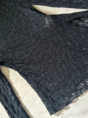 Svart spetsblus med lång ärm - Säljer en svart blus i genombruten spets med lång ärm. Toppen har en figurnära passform och är helsvart med vackert spetsmönster över hela plagget. Perfekt att styla lager på lager eller med ett linne under.
