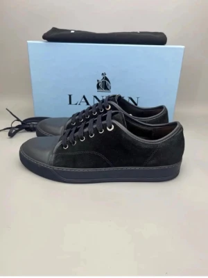 Svarta sneakers från Lanvin - Stilrena svarta sneakers från Lanvin med ovandel i mocka och detaljer i slätt läder. Skorna har svarta skosnören, metallfärgade öljetter och en svart sula. Perfekta för dig som gillar en clean och klassisk look.