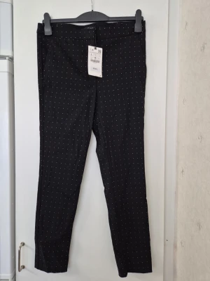 Kostymbyxor - Säljer ett par svarta kostymbyxor med små vita prickar och smal passform. Byxorna har klassisk midja, bälteshällor och diskreta fickor. Perfekta för dig som vill ha en stilren men ändå lekfull look. Men känns mer som M, beror på midjestorlek vad man har. Nypris: 345kr