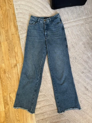 Blå raka jeans med rå kant - Säljer ett par blå raka jeans med hög midja och rå, fransig kant längst ner på benen. Jeansen har klassisk femficksdesign och är gjorda i ett kraftigt denimtyg. Perfekta för en avslappnad och trendig look.