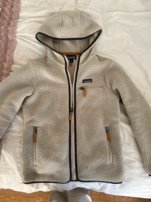 Beige fleecejacka från Patagonia - Mysig beige fleecejacka från Patagonia med huva och dragkedja framtill. Jackan har tre fickor med dragkedja och mörkblå detaljer längs kanterna. Perfekt nu till vår och sommar. Nästan aldrig använd och tvättas innan den skickas. Pris kan diskuteras