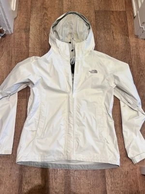 Vit regnjacka från The North Face - Snygg vit regnjacka från The North Face med huva och dragkedja framtill. Jackan har två sidofickor med dragkedja och justerbar huva. Tillverkad i ett vattenavvisande material som håller dig torr under regniga dagar. Perfekt för dig som vill ha en stilren och funktionell jacka.