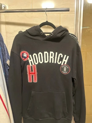 Svart hoodie från Hoodrich  - Svart hoodie från Hoodrich med stor vit logga på bröstet, röda och vita patches samt en stor ficka framtill. Det är ett litet märke på luvan som ej går bort tyvärr.