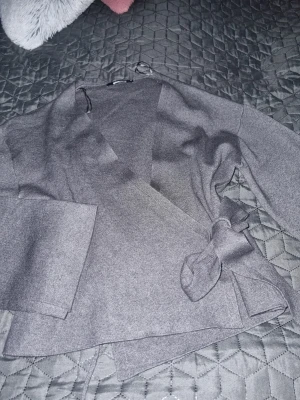 Grå omlottkofta med knytband från Ginatricot  - Snygg grå omlottkofta med breda ärmar och knytband i midjan. Koftan har en avslappnad passform och är tillverkad i mjukt stickat material. Perfekt att slänga över en topp för en chill look. Bara provat I storlek M. Ny utan prislapp 