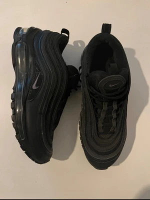 Nike Air Max 97 Triple Black sneakers - Snygga Nike Air Max 97 Triple Black sneakers med vågformad design och synlig Air-sula. Helt svarta med diskret Swoosh på sidan och mesh- samt syntetmaterial. Perfekta för dig som gillar streetwear och sportig stil.