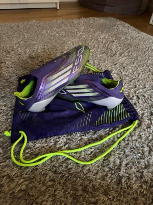 Adidas F50 lila fotbollsskor - Säljer ett par Adidas F50 fotbollsskor i lila med vita och neongula detaljer. Skorna har en strömlinjeformad design och levereras med en matchande lila och neongul skopåse. Perfekta för dig som vill sticka ut på planen.