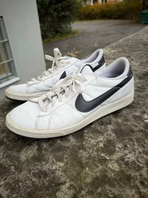 Vita Nike sneakers med svart swoosh - Klassiska vita sneakers från Nike med svart swoosh på sidorna. Skorna har lågt skaft, vita snören och ovandel i läder. Yttersulan är vit med en lätt textur. Perfekta för en clean och sportig stil. 45,5