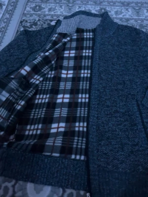 Mörkgrå ziptröja med rutigt foder - Mysig mörkgrå ziptröja med ståkrage och ribbade muddar. Insidan har ett mjukt rutigt foder i vitt, brunt och svart som ger extra värme. Två fickor framtill och grovstickad känsla. Perfekt för kyliga dagar.