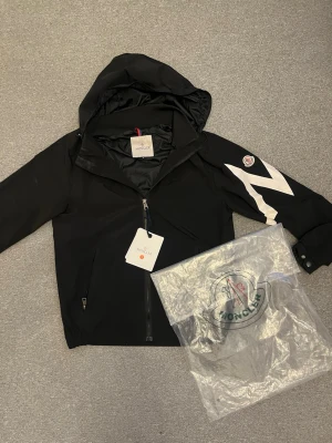 Svart Moncler vindjacka med huva - Snygg svart vindjacka från Moncler med stor vit blixtdetalj på ärmen och Moncler-logga på axeln. Jackan har huva, dragkedja framtill och justerbara muddar. Tillverkad i ett lätt och tåligt material, perfekt för blåsiga dagar.