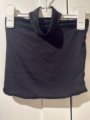 Svart tubhalsduk i trikå - En enkel och stilren svart tubhalsduk i mjuk trikå. Halsduken har en rundad form och är lätt att dra över huvudet. Perfekt för att hålla värmen under kyliga dagar och passar till de flesta outfits.