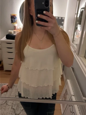Vit volangtop med tunna axelband - Söt vit topp med flera lager volanger och tunna axelband. Toppen har en luftig och flowy känsla, perfekt för en trendig och avslappnad look. Passar snyggt till både jeans och kjol.