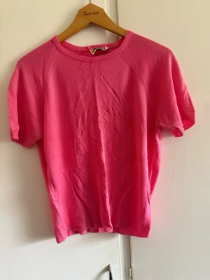  Vintage 1950 Rosa stickad kortärmad topp från Queenesta by Rowley  - A vintage rosa stickad topp med korta ärmar och rund halsringning. Toppen har ett diskret mönster framtill som ger en extra detalj. Perfekt att styla med jeans eller kjol för en fräsch look.