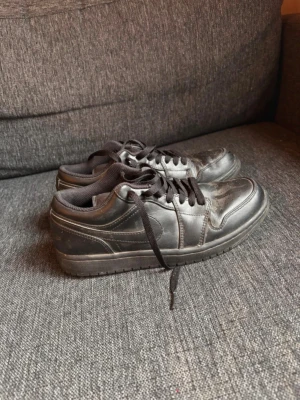 Svarta Nike Air Jordan 1 Low sneakers - Svarta Nike Air Jordan 1 Low sneakers i helsvart läder med matchande svarta skosnören och diskret broderad Air Jordan-logga på hälen. Klassisk siluett med lågt skaft och vadderad plös för extra komfort. Perfekt för dig som gillar streetwear och tidlös stil.