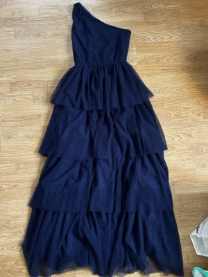 Balklänning  - Marinblå långklänning med flera lager volanger och oneshoulder-design. Klänningen har en snygg rosettdetalj på axeln. Jag hade denna på min bal. Den är insydd som storlek 34 men går jättelätt att sprätta upp till 36