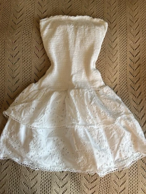 Vit axelbandslös kort klänning  - Söt vit axelbandslös kort klänning med smockad överdel och volangig kjol i broderad bomull. Kjolen har fina spetsdetaljer och blommigt broderi som ger en romantisk känsla. Perfekt för varma dagar och somriga tillfällen.