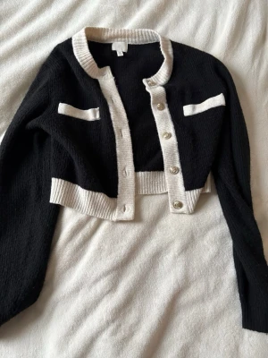 Svart och vit cropped kofta från H&M - Trendig svart cropped kofta från H&M med vita ribbade kanter och dekorativa fickor framtill. Koftan har långa ärmar, rund halsringning och snygga knappar i storlek S